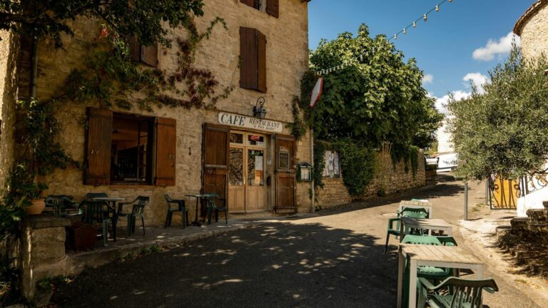 Café pittoresque en pierre en Provence avec des volets en bois, des tables extérieures et de la verdure, situé dans une rue calme sous un ciel bleu clair.