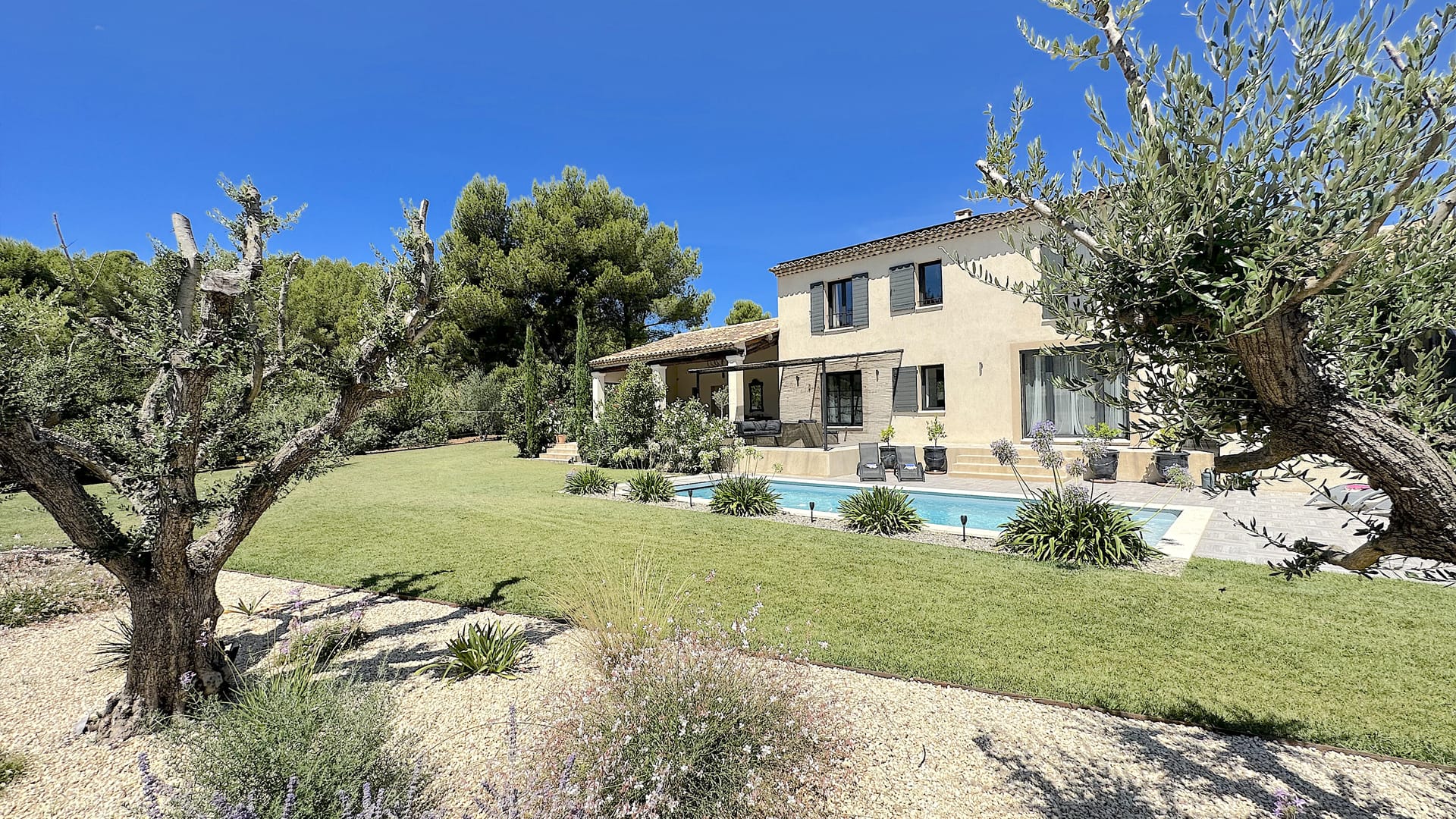 Villa provençale entourée de jardins verdoyants, piscine bordée de plantes et oliviers sous un ciel bleu éclatant.