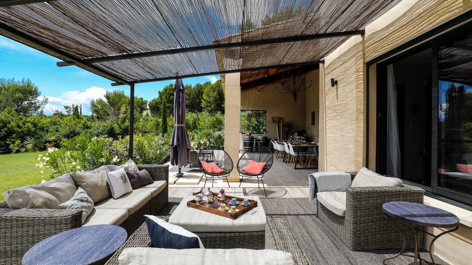 Terrasse extérieure avec mobilier en osier et coussins, espace couvert et table avec rafraîchissements. Une végétation luxuriante entoure l'espace sous un ciel bleu clair.